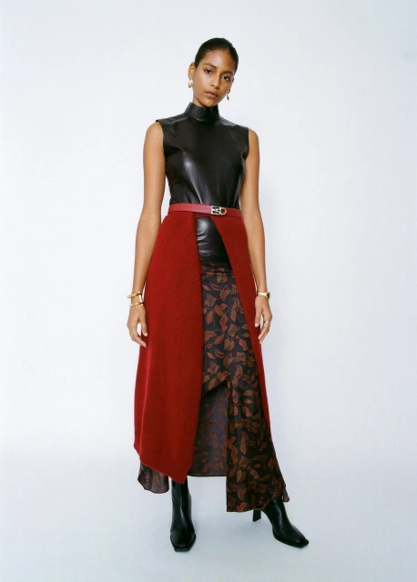 A woman poses in a sleeveless black top and red skirt in a studio shot, accessorized with gold jewelry and black boots.