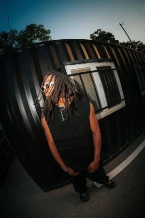 A young adult with dreadlocks in black sleeveless top and pants stands outdoors next to a shipping container, wearing round sunglasses.