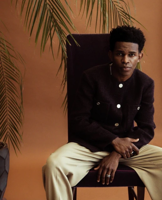 Stylish portrait of a handsome Black man seated indoors with palms and a textured jacket against a warm brown backdrop, relaxed pose.