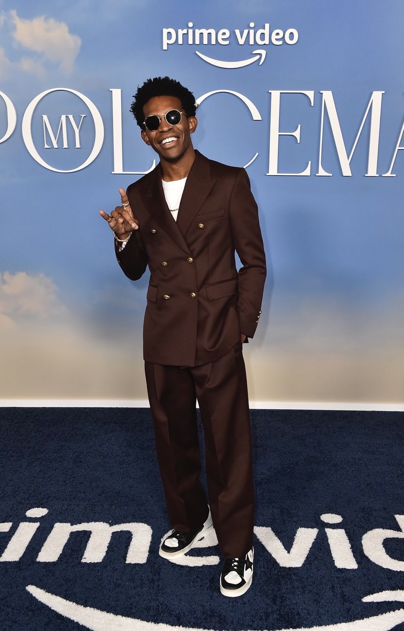 A young Black adult poses in a brown suit on a red carpet with the "Prime Video" logo, smiling, with sunglasses and a hand gesture.