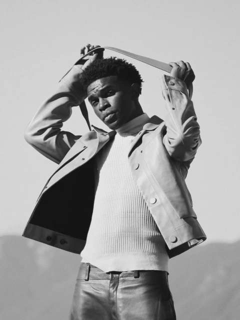 Monochrome portrait of a Black male wearing a cream ribbed sweater, light jacket, and leather pants, holding a strap above his head.