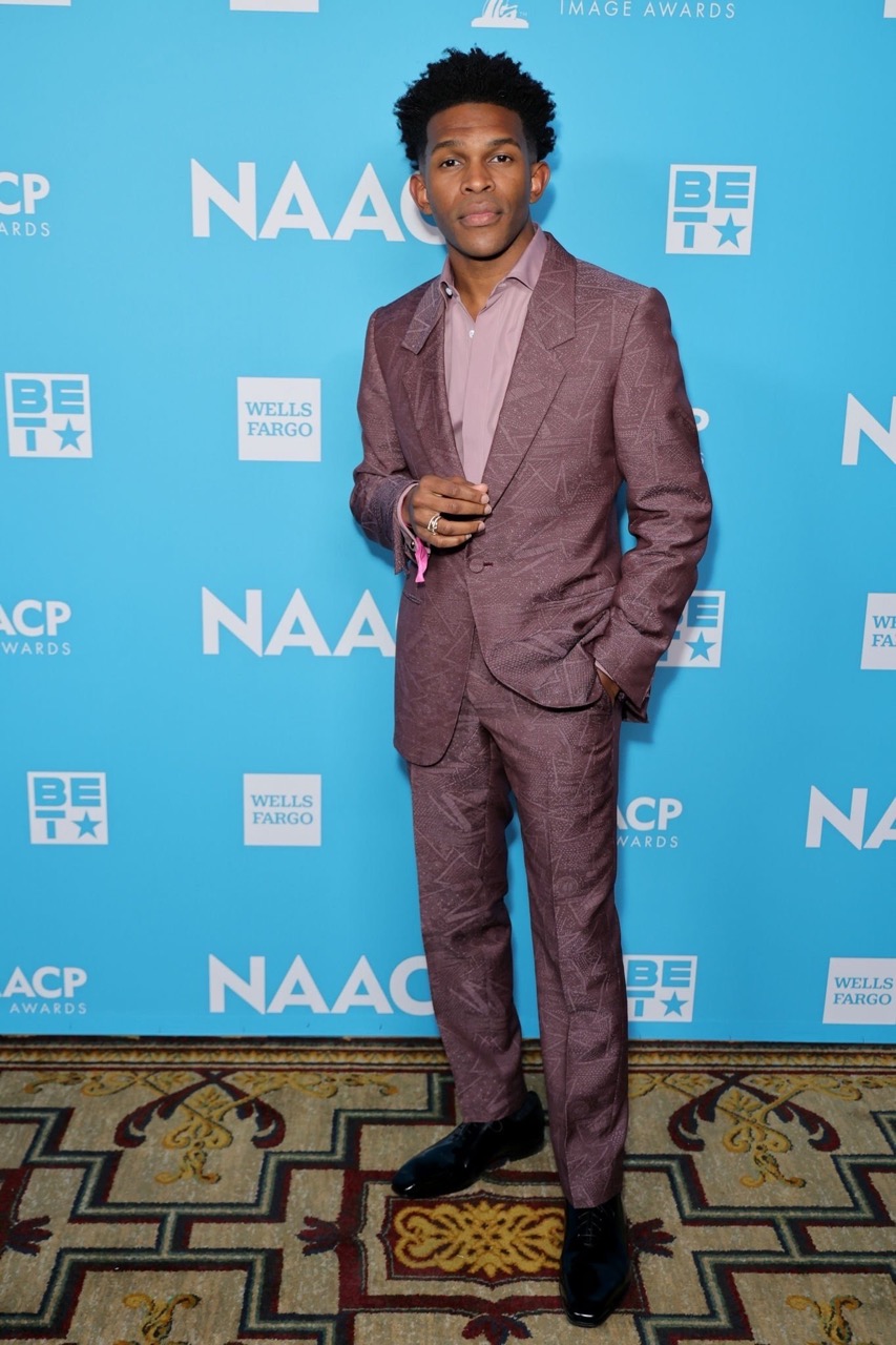 A confident Black adult male in a mauve suit stands against a branded backdrop, his hands clasped, radiating professionalism and poise.