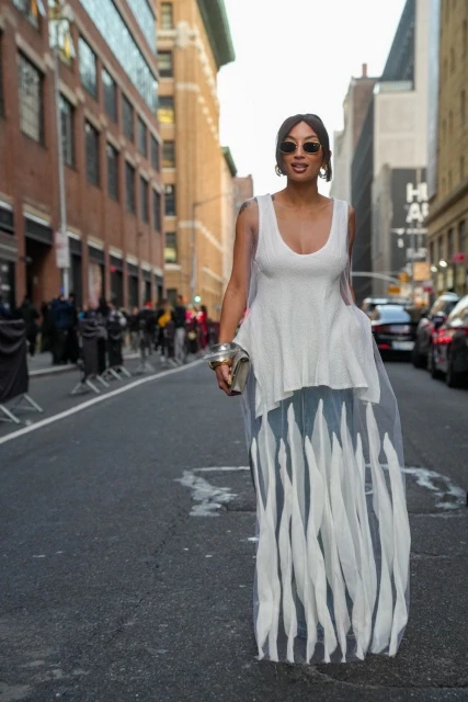 Woman in flowing white dress walks confidently down a city street, accessorized with sunglasses and silver bangle, under natural daylight.