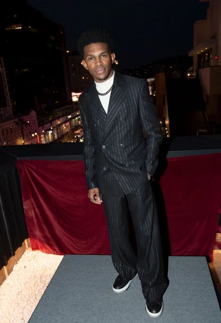 An African American man poses in a pinstripe suit against a dark urban backdrop, exhibiting an editorial fashion style at nighttime.
