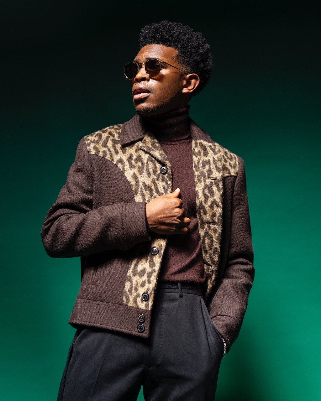 Black man wearing sunglasses, brown leopard-print panel jacket, and dark trousers against a green background.