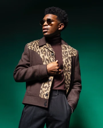 Black man wearing sunglasses, brown leopard-print panel jacket, and dark trousers against a green background.