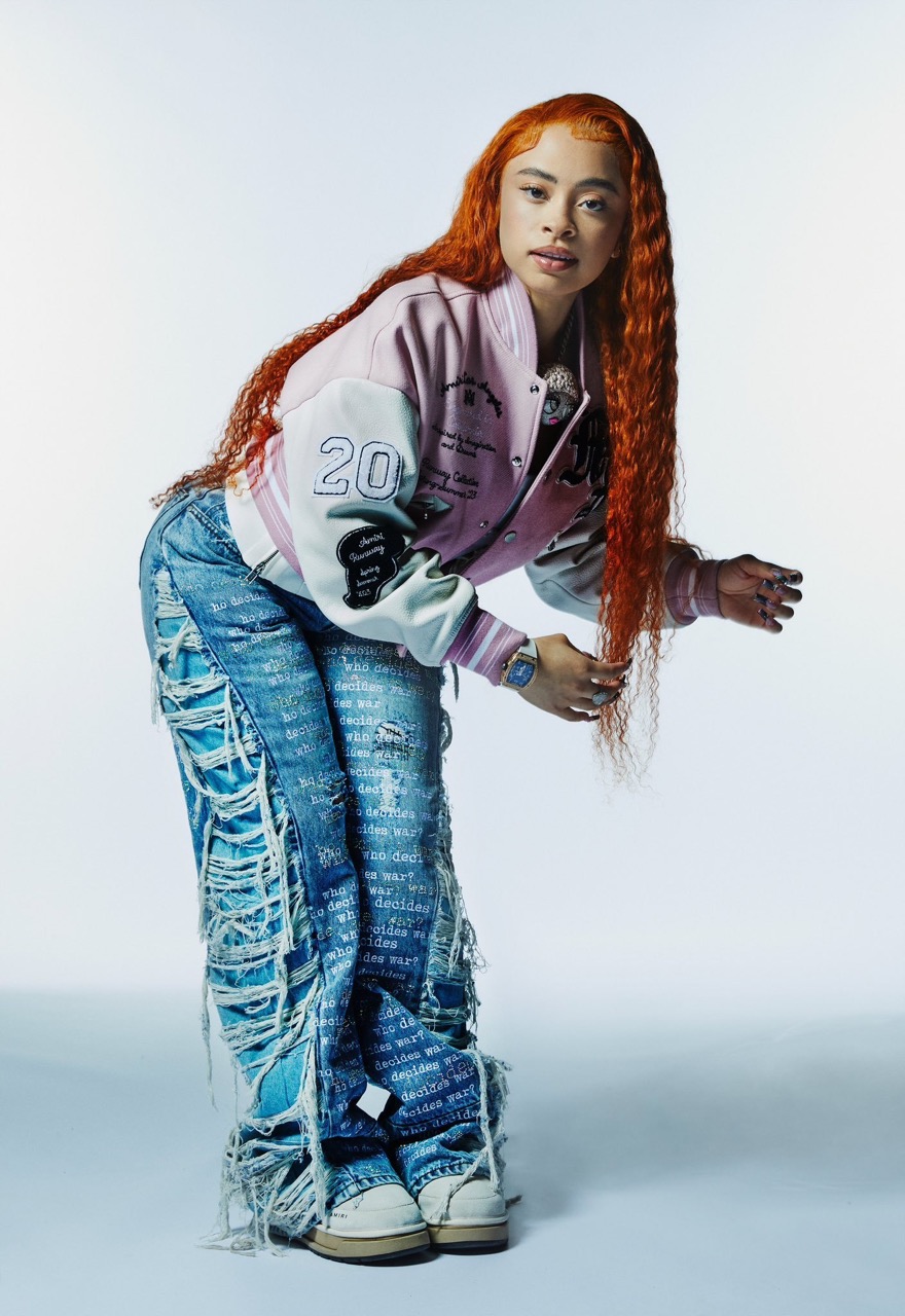 Model with long curly red hair poses in an unbuttoned light pink and white letterman jacket and distressed denim pants in a studio.