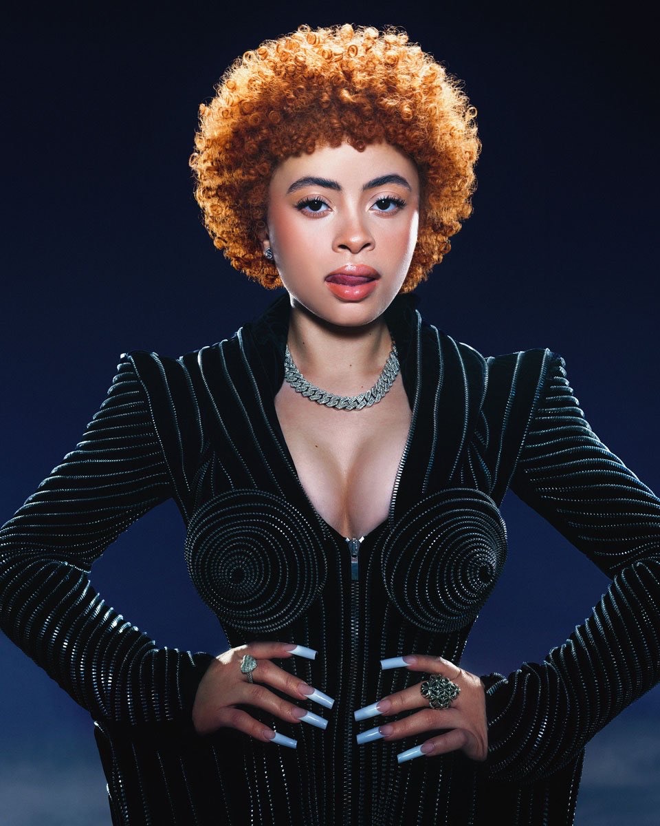Portrait of a young woman with orange hair, wearing a sparkling black jacket and jewelry, posing against a dark blue background.