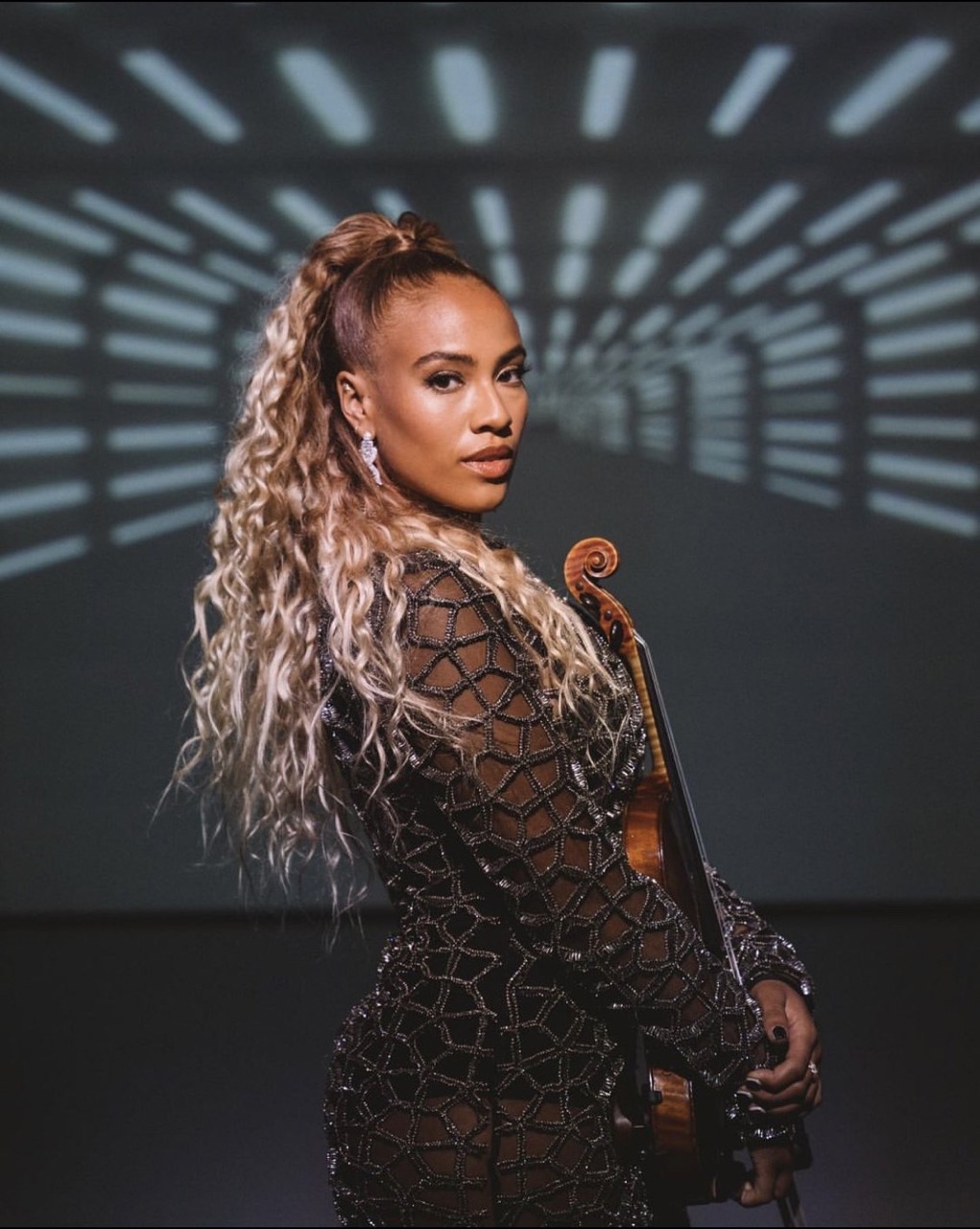 Female musician with a violin, wearing a long-sleeved patterned dress and earrings, with curly hair in a ponytail, in front of a light display.