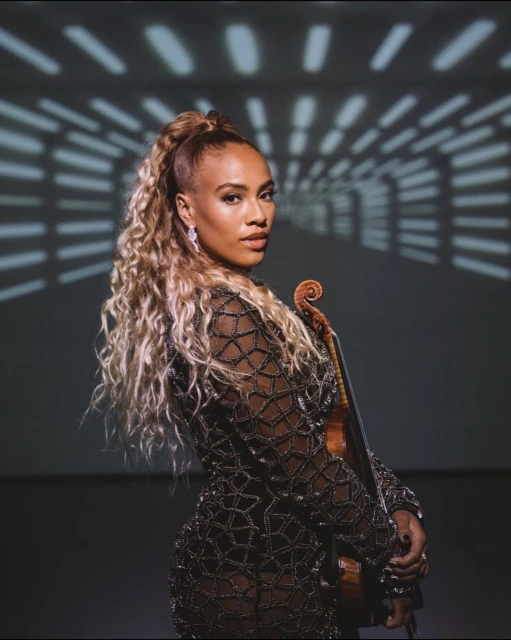 Female musician with a violin, wearing a long-sleeved patterned dress and earrings, with curly hair in a ponytail, in front of a light display.