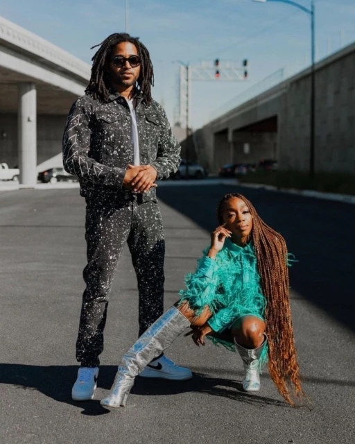 A stylish couple poses on a road under an overpass; the woman wears silver boots and the man is in a splattered denim suit.