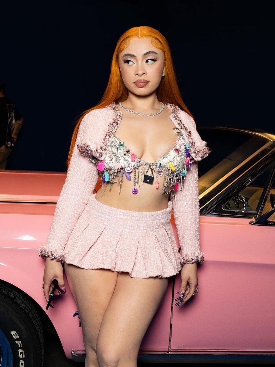 Woman with orange hair in a pink tweed set stands next to a pink classic car in an indoor studio against a dark background.