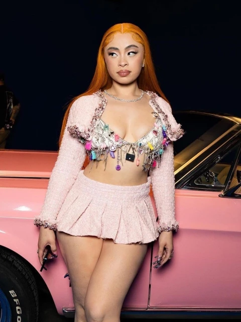 Woman with orange hair in a pink tweed set stands next to a pink classic car in an indoor studio against a dark background.