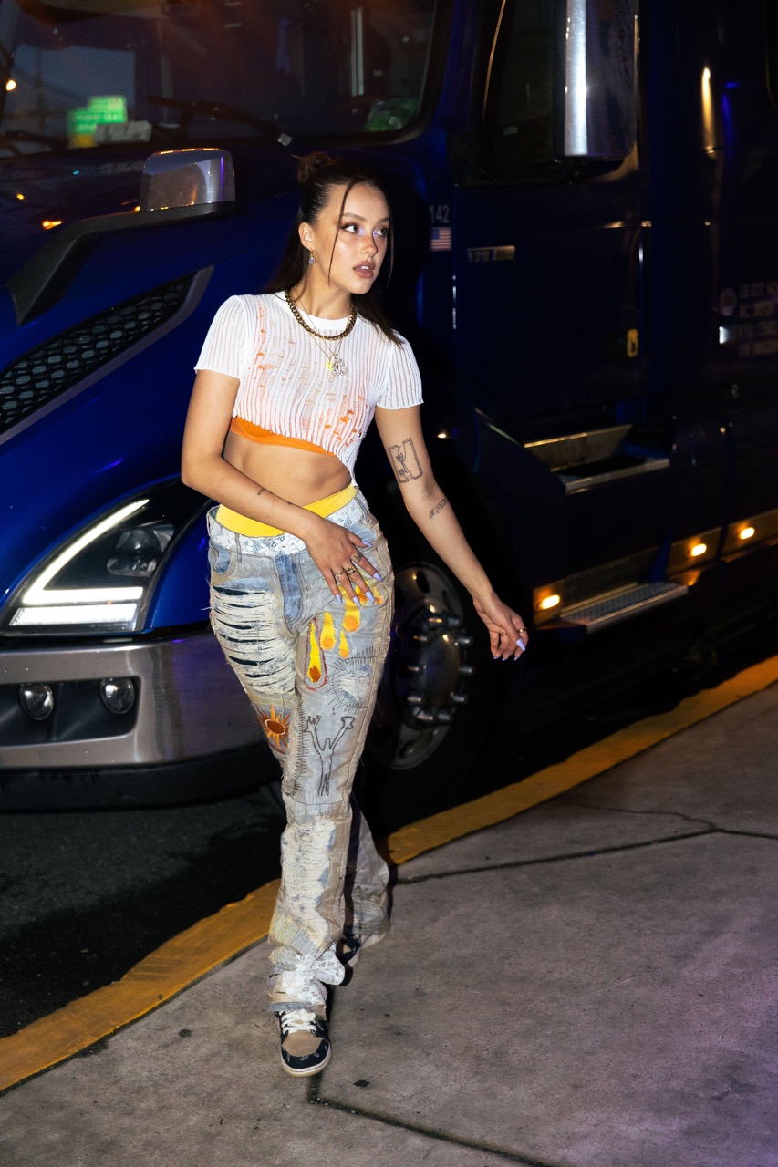 A young woman poses in front of a semi-truck wearing a white crop top and painted denim jeans in an outdoor urban setting.