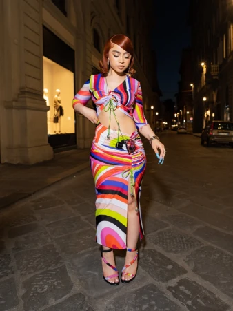 A stylish woman with red hair poses on a city street, wearing a colorful patterned two-piece outfit and strappy heels at dusk.