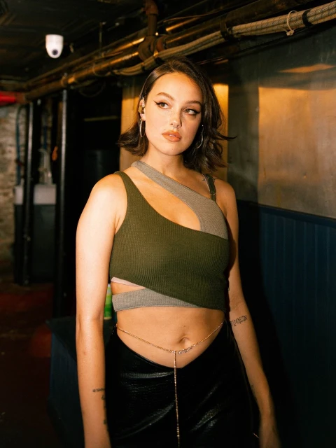Portrait of woman in a green and grey cut-out top and black pants posing indoors with an exposed belly chain.