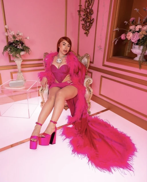 Adult female model in pink corset dress, fur robe and platform heels poses on a gold throne in a pink photo studio.
