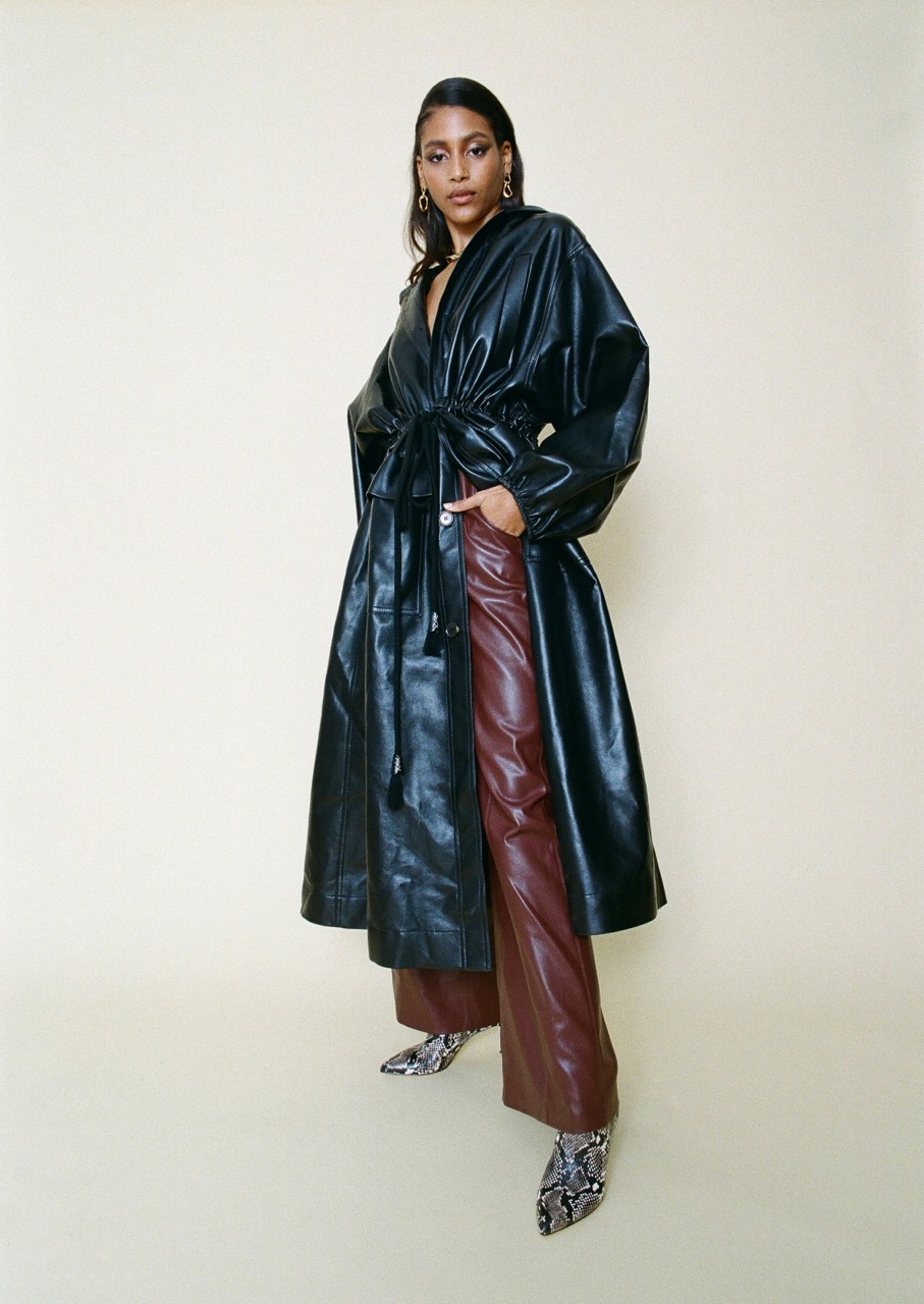 A stylish African-American woman poses in a long, black, belted, faux-leather trench coat, brown leather pants, and snakeskin ankle boots against a cream backdrop.