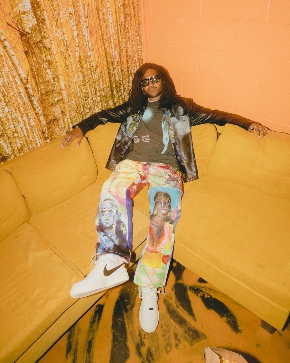 An adult male with long dark hair is sitting on a yellow couch wearing a brown t-shirt, jacket and unique jeans and white sneakers.