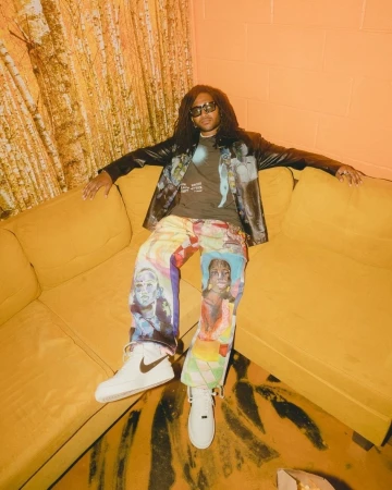 An adult male with long dark hair is sitting on a yellow couch wearing a brown t-shirt, jacket and unique jeans and white sneakers.