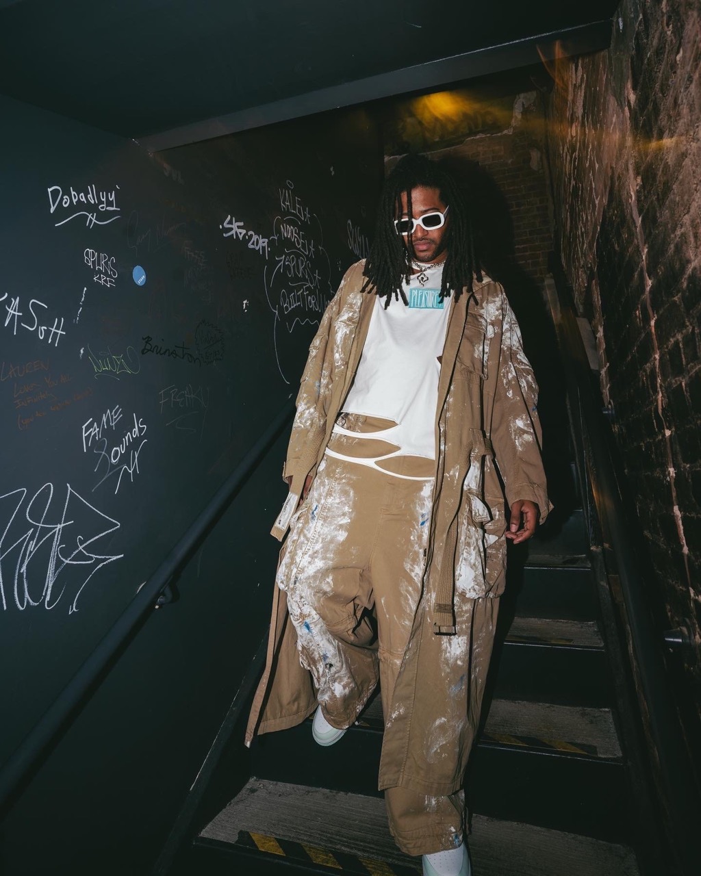 Person with dreadlocks descends stairs in tan painter pants and trench coat, wearing sunglasses, captured in a gritty, urban environment.