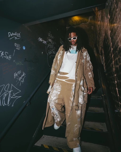 Person with dreadlocks descends stairs in tan painter pants and trench coat, wearing sunglasses, captured in a gritty, urban environment.