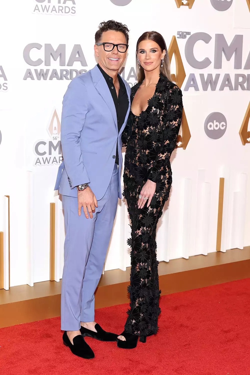 Man and woman pose on red carpet at the CMA Awards. The man wears a blue suit, while the woman is in a long black lace dress.