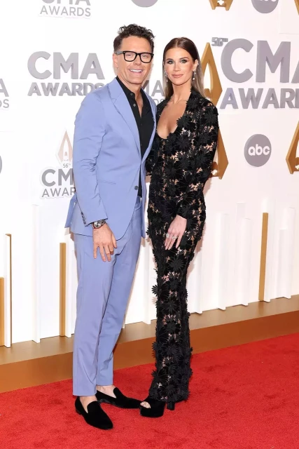 Man and woman pose on red carpet at the CMA Awards. The man wears a blue suit, while the woman is in a long black lace dress.