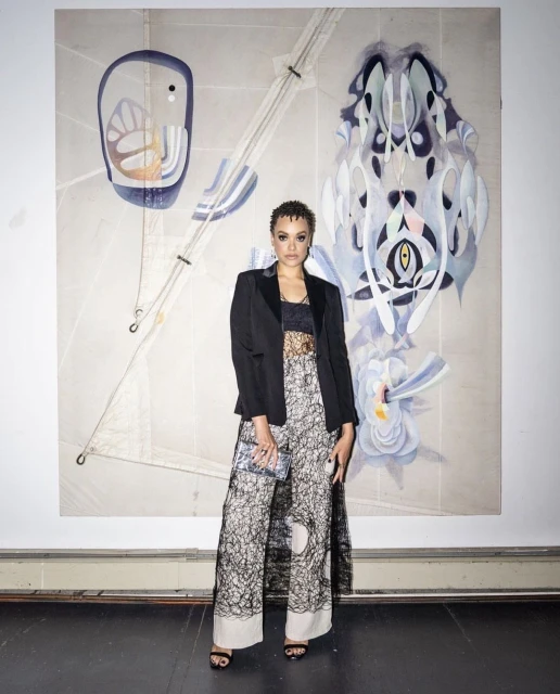 Woman poses in black blazer and patterned pantsuit in front of large-scale abstract art. Indoor environment, dark flooring.
