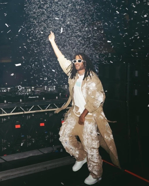DJ with dreadlocks and white sunglasses performing on stage under confetti. Man wears paint-splattered clothing and white sneakers.