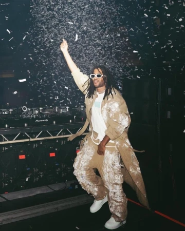 DJ with dreadlocks and white sunglasses performing on stage under confetti. Man wears paint-splattered clothing and white sneakers.