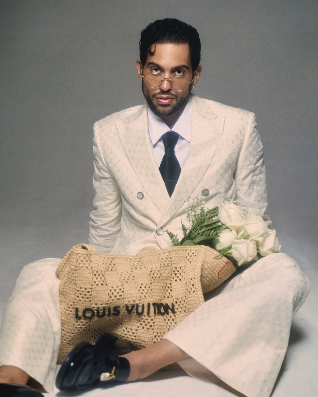 Adult male in a double-breasted patterned suit sitting on a gray background with a designer bag and bouquet of white roses.