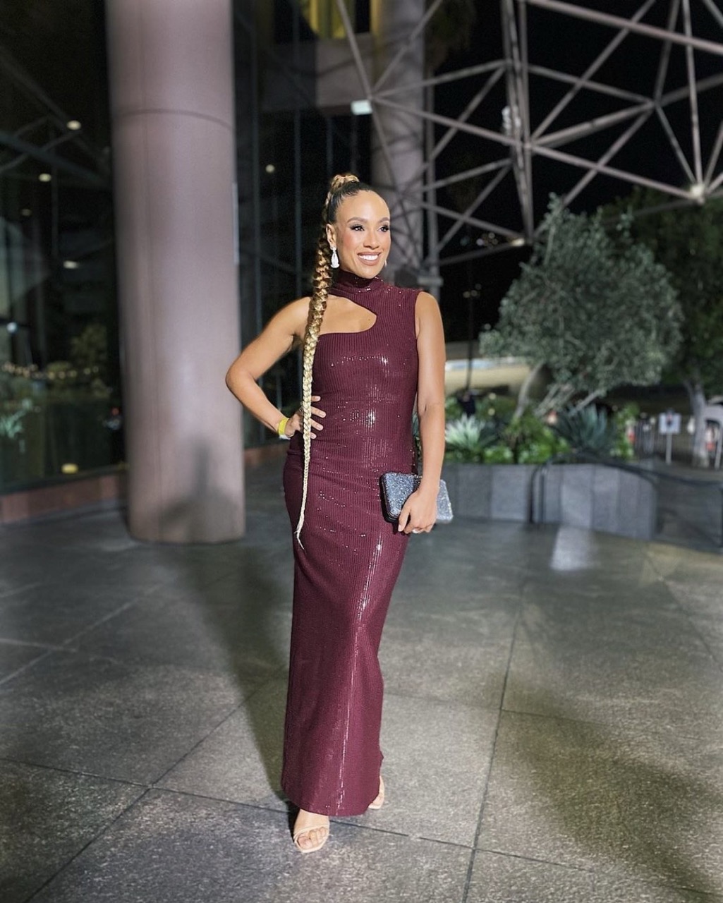Smiling woman in maroon sequin gown with a cutout poses outdoors at night with buildings and greenery in the background.