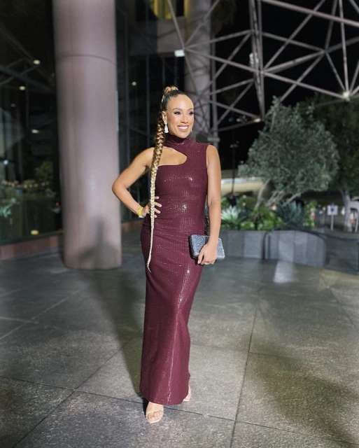 Smiling woman in maroon sequin gown with a cutout poses outdoors at night with buildings and greenery in the background.
