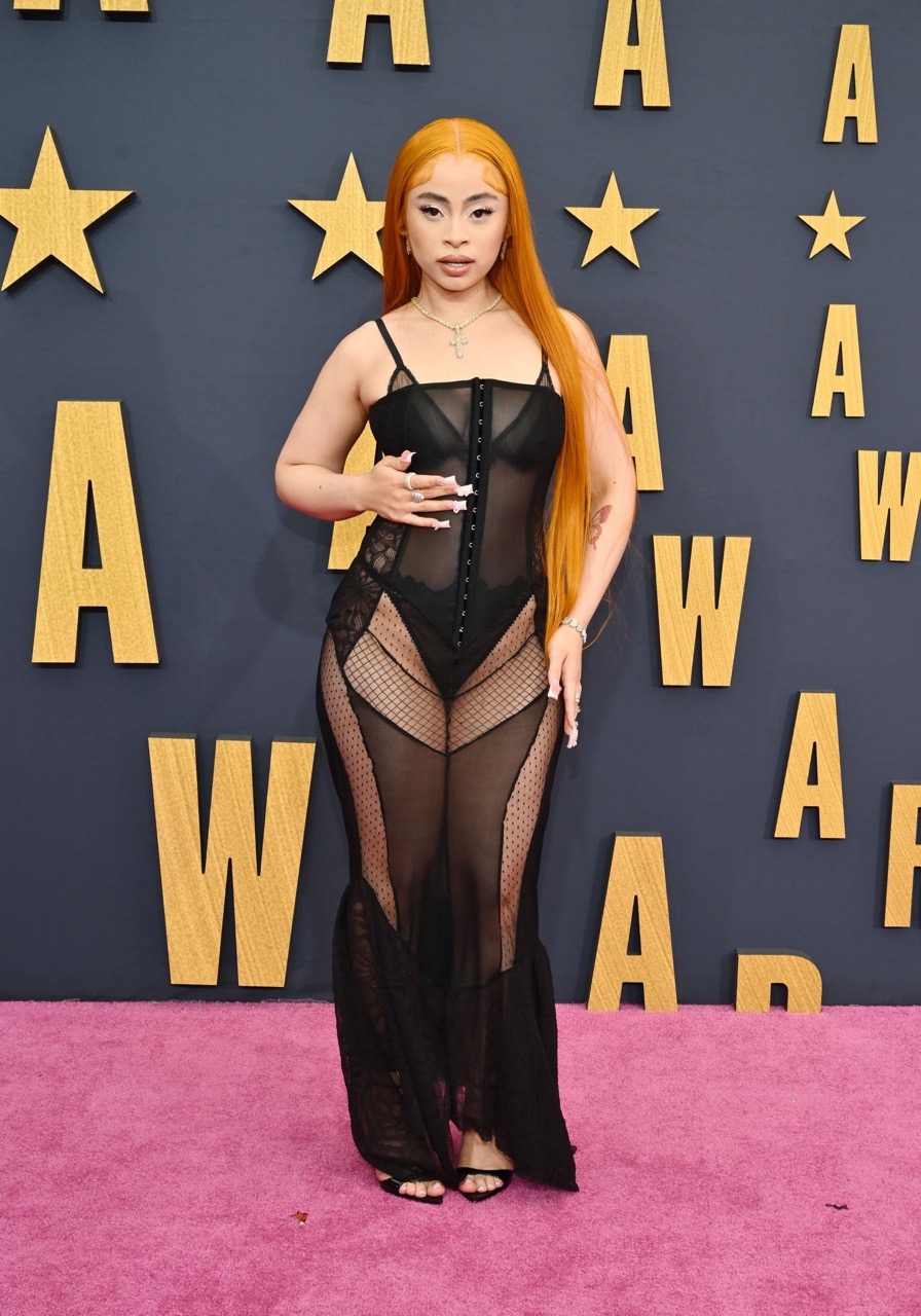 Woman with long orange hair in sheer black dress on a pink carpet at an awards show with a star-studded wall. Full-length shot.