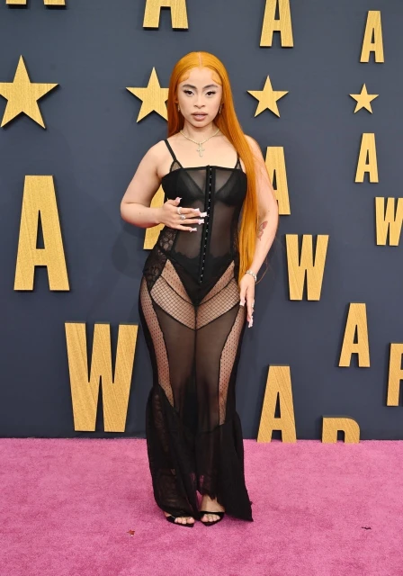 Woman with long orange hair in sheer black dress on a pink carpet at an awards show with a star-studded wall. Full-length shot.