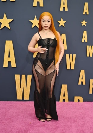 Woman with long orange hair in sheer black dress on a pink carpet at an awards show with a star-studded wall. Full-length shot.