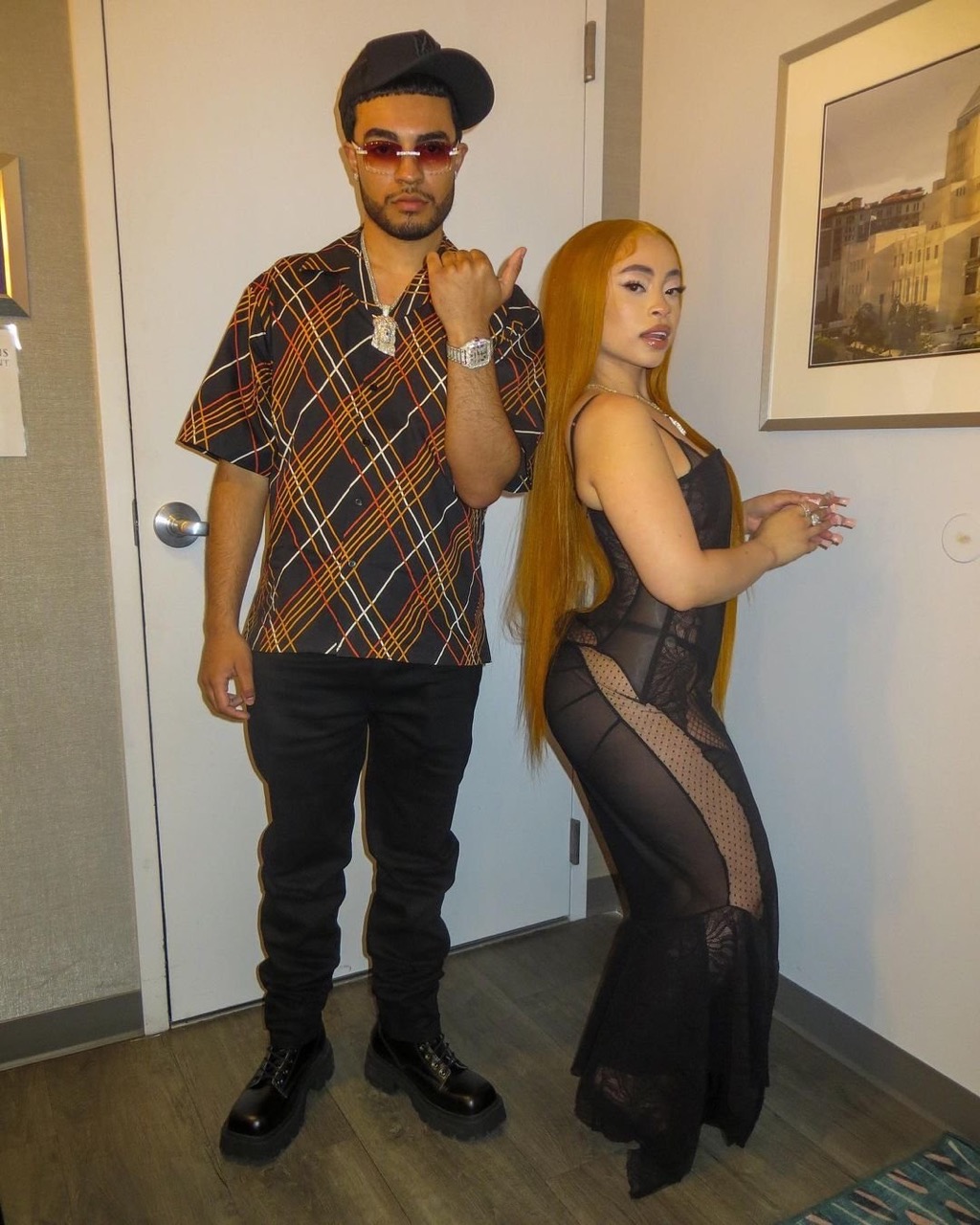 A stylishly dressed couple poses indoors against a neutral background. The man wears a patterned shirt and the woman a black dress.