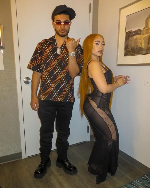A stylishly dressed couple poses indoors against a neutral background. The man wears a patterned shirt and the woman a black dress.