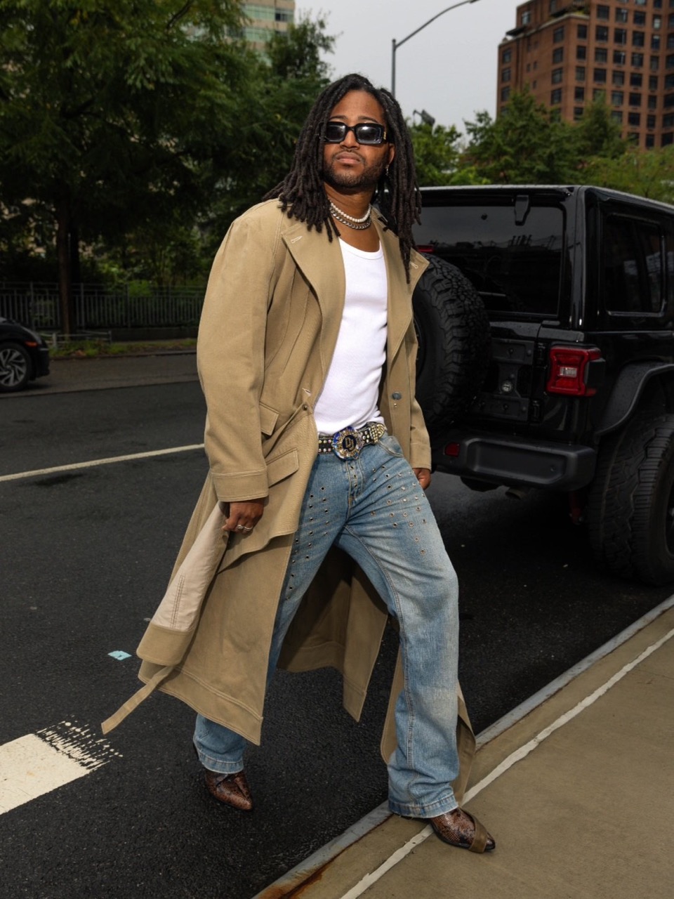 An adult male with locs wears a tan trench coat, jeans, and snakeskin shoes, posing on a city street next to a black SUV with a spare tire.