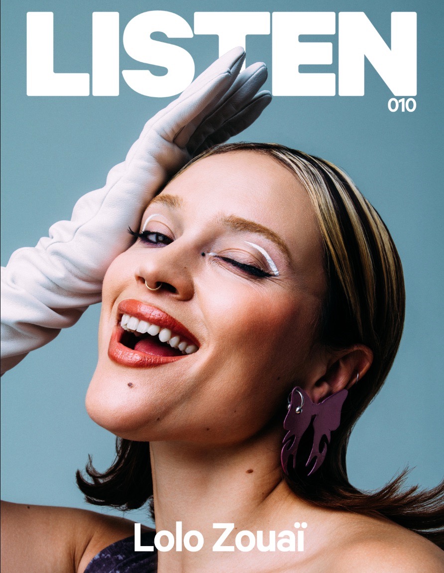 Magazine cover featuring a smiling adult woman with creative makeup, purple statement earrings, wearing a white glove, against a light blue backdrop.