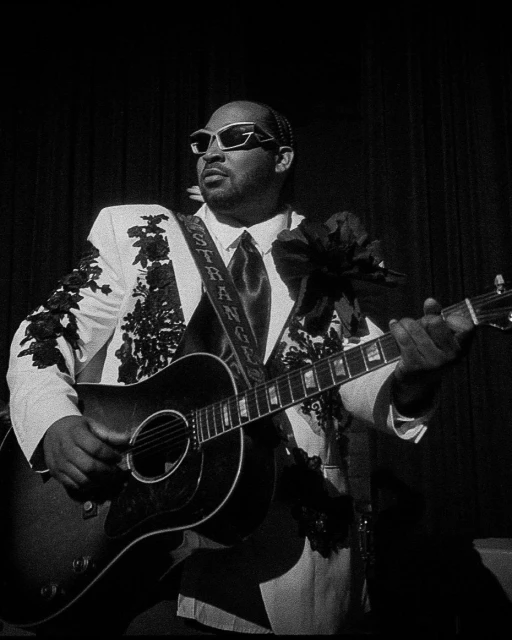 Monochrome portrait of an adult male musician in an embellished white suit playing an acoustic guitar on a dimly lit stage.