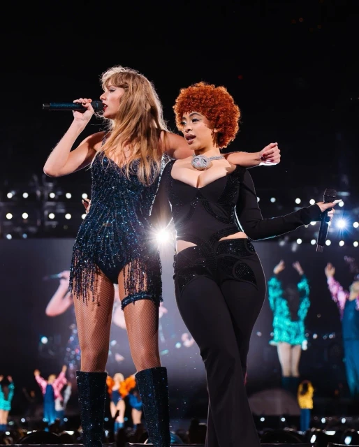 Taylor Swift and Ice Spice perform on stage at a concert, wearing embellished costumes and holding microphones, with a blurred crowd.