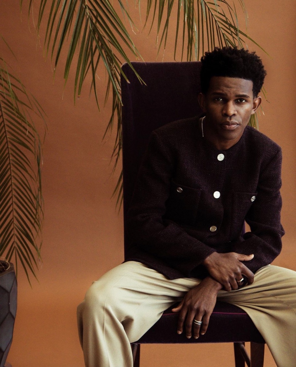Portrait of a young adult male with dark brown skin sitting on a chair in front of a brown backdrop with tropical leaves.