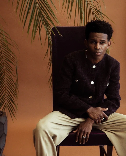 Portrait of a young adult male with dark brown skin sitting on a chair in front of a brown backdrop with tropical leaves.