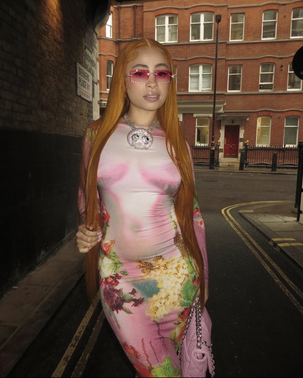 Woman with long orange hair wearing pink tinted sunglasses and floral print dress standing on a street in front of a red brick building.