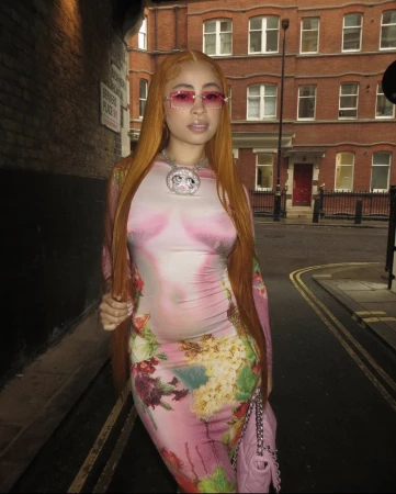 Woman with long orange hair wearing pink tinted sunglasses and floral print dress standing on a street in front of a red brick building.