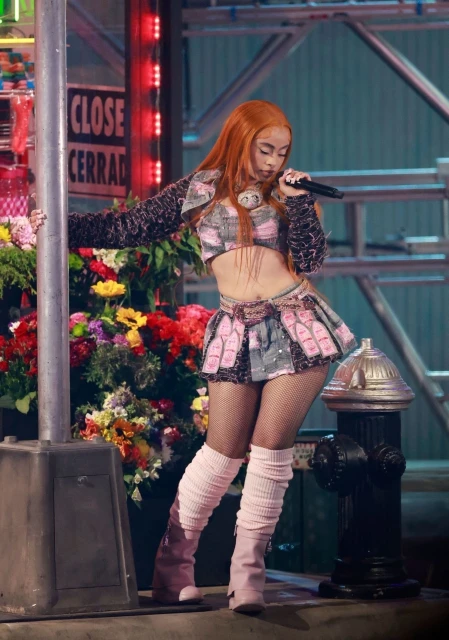 Singer in a cropped top and skirt performs on stage with a microphone, flowers, and a fake building facade in the background.