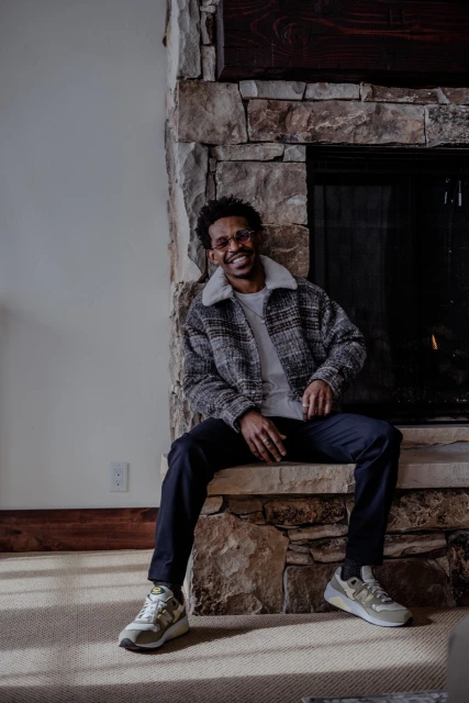 A smiling African American man sits in front of a stone fireplace wearing a jacket, pants, and sneakers in an indoor setting.