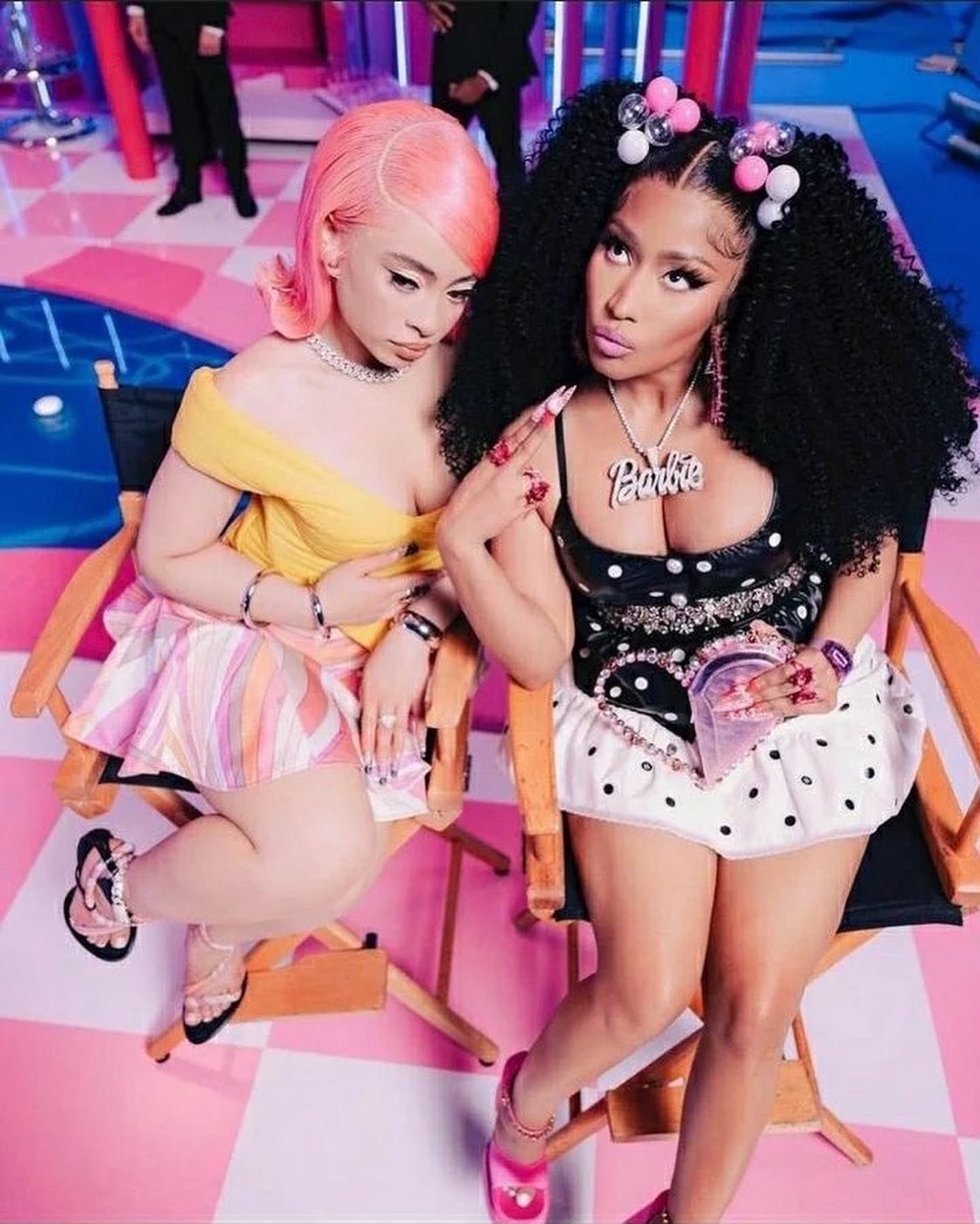 Two women, one with pink hair and the other with black hair and a Barbie necklace, pose in a photo studio with a checkered floor.